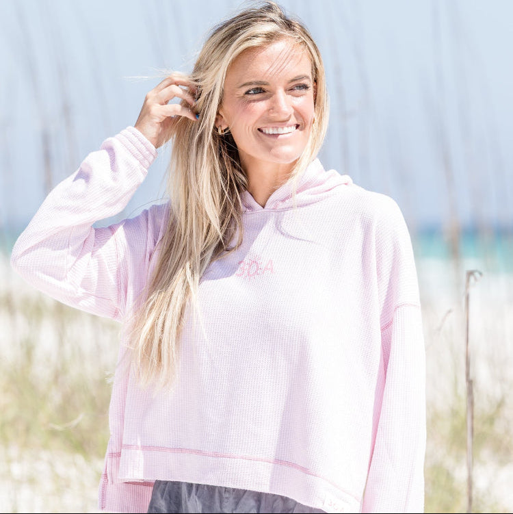 Woman wearing a light pink hoodie with a logo, standing outdoors with a blurred background