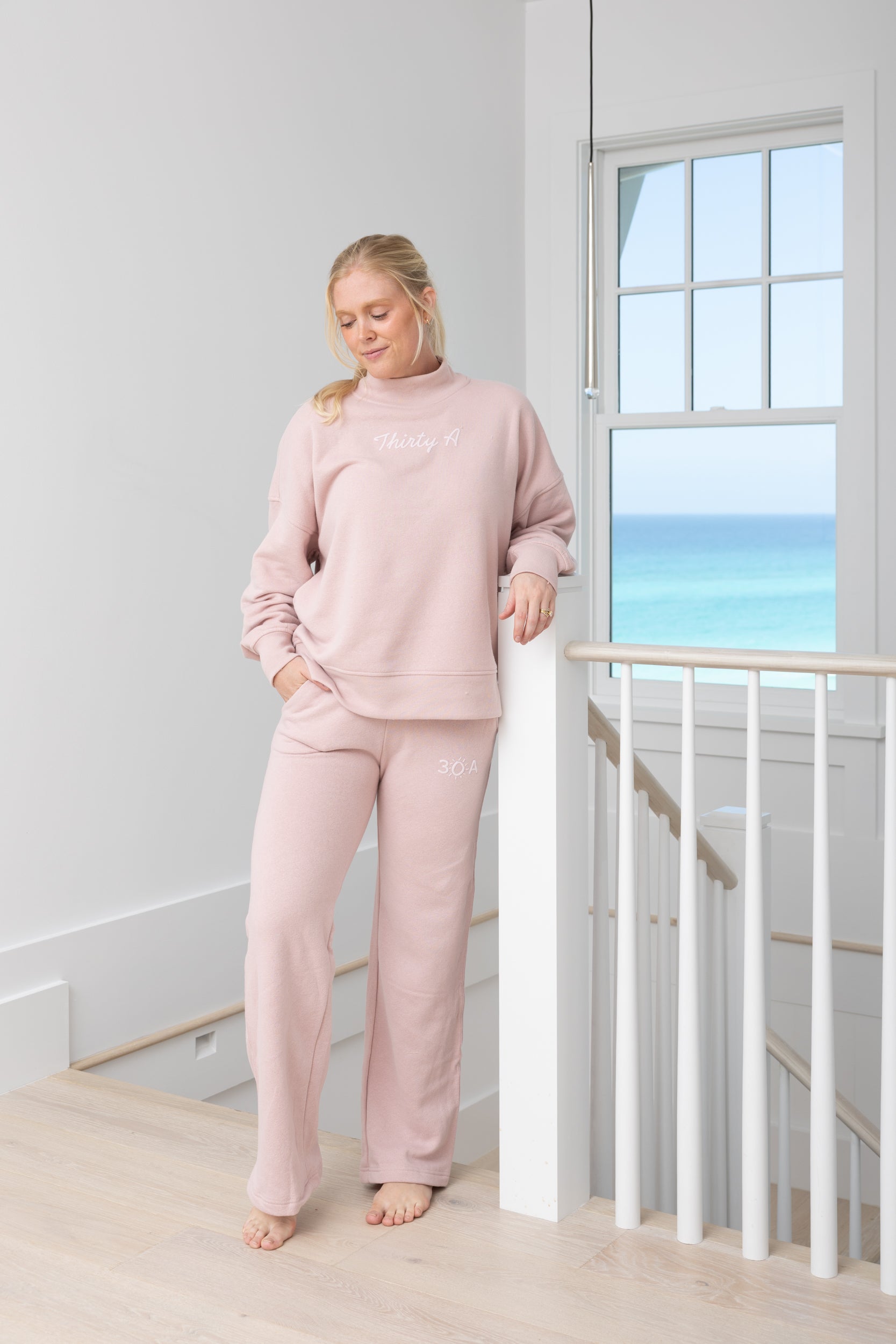 Woman wearing a pink loungewear set standing by a window with a view of the ocean.