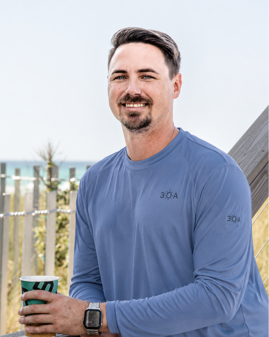 Man wearing a blue long-sleeve shirt with '30A' logo, holding a cup, standing on a wooden deck with beach view.